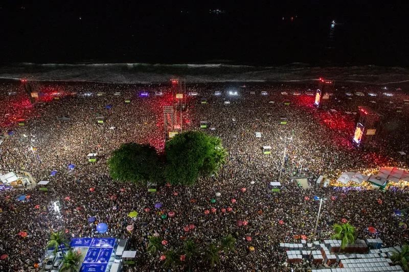 A imagem captura uma multidão imensa reunida na Praia de Copacabana durante o show de abertura da Lady Gaga, em um evento noturno vibrante. As pessoas estão aglomeradas, criando um mar de cabeças que se estende até onde a vista alcança, com algumas luzes de palco iluminando a cena. O ambiente é festivo, com uma mistura de cores das roupas e dos acessórios dos participantes, além de tendas e estruturas de palco visíveis ao fundo.